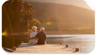 An elderly couple leisurely sitting on a bench near a lake enjoying their sunset.